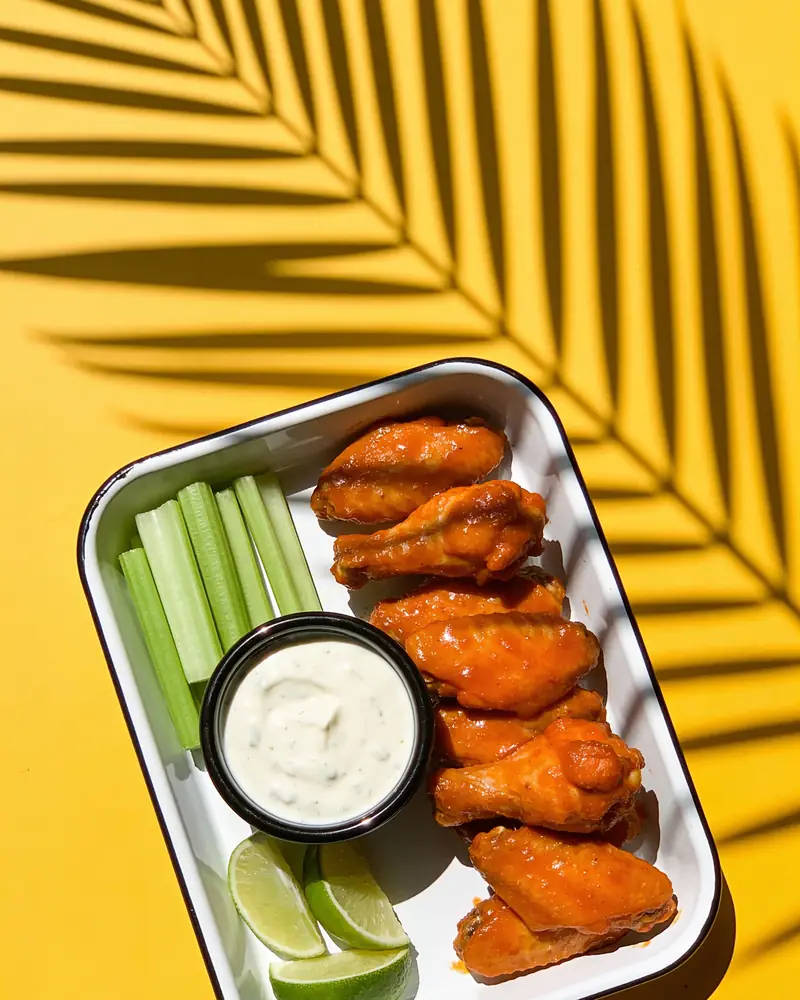 Buffalo wings with ranch and celery on a yellow table with palm shadows